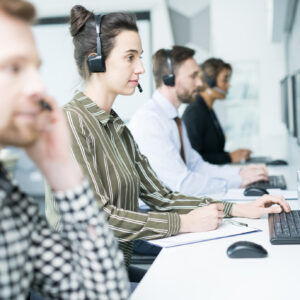 Customer Service Operator Side view portrait of several help desk operators wearing headsets sitting in row looking at computer screens and talking to clients